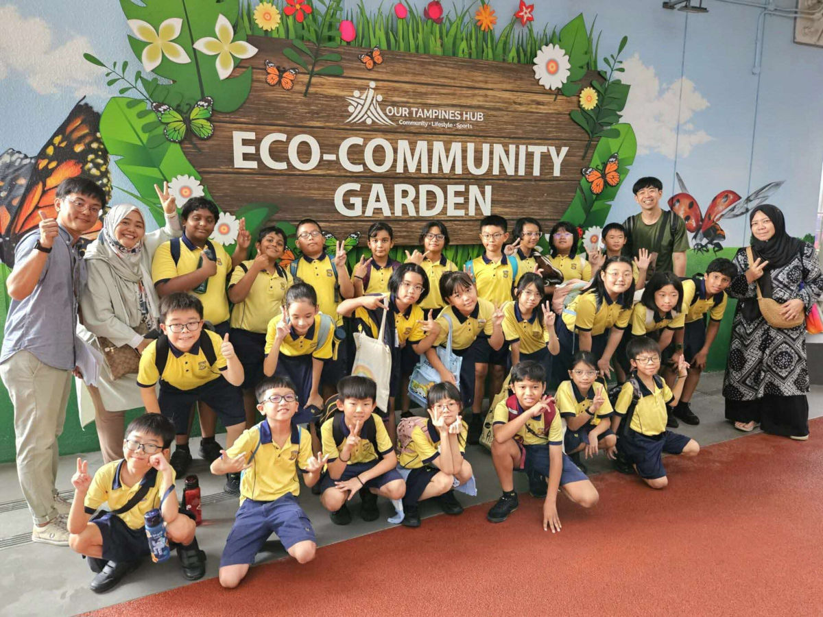 A group of children smiling and posing together in front of a vibrant eco community garden filled with plants and flowers